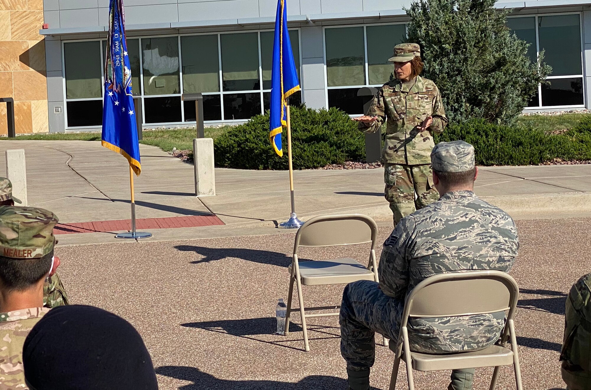 A woman stand outside near two flags speaking to an audience.