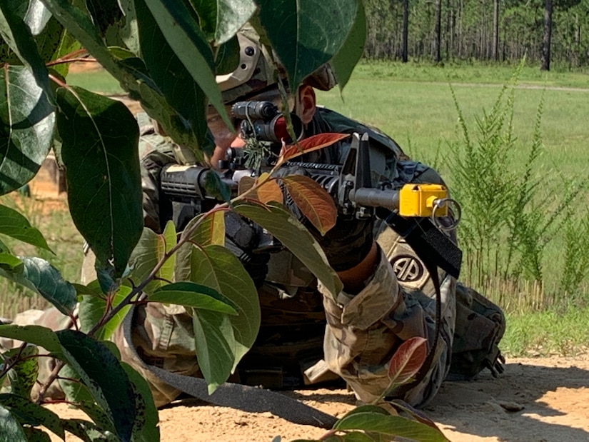 A soldier lying prone and wearing a camouflage uniform aims a rifle while using vegetation as cover.