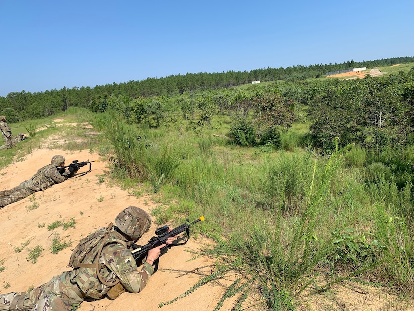 Soldiers wearing camouflage uniforms aim their rifles as they lie prone at the crest of a slope at the edge of an area covered with vegetation.