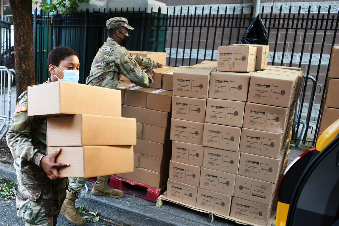 A guardsman wearing a face mask carries boxes of meals to a vehicle for distribution at a food site.