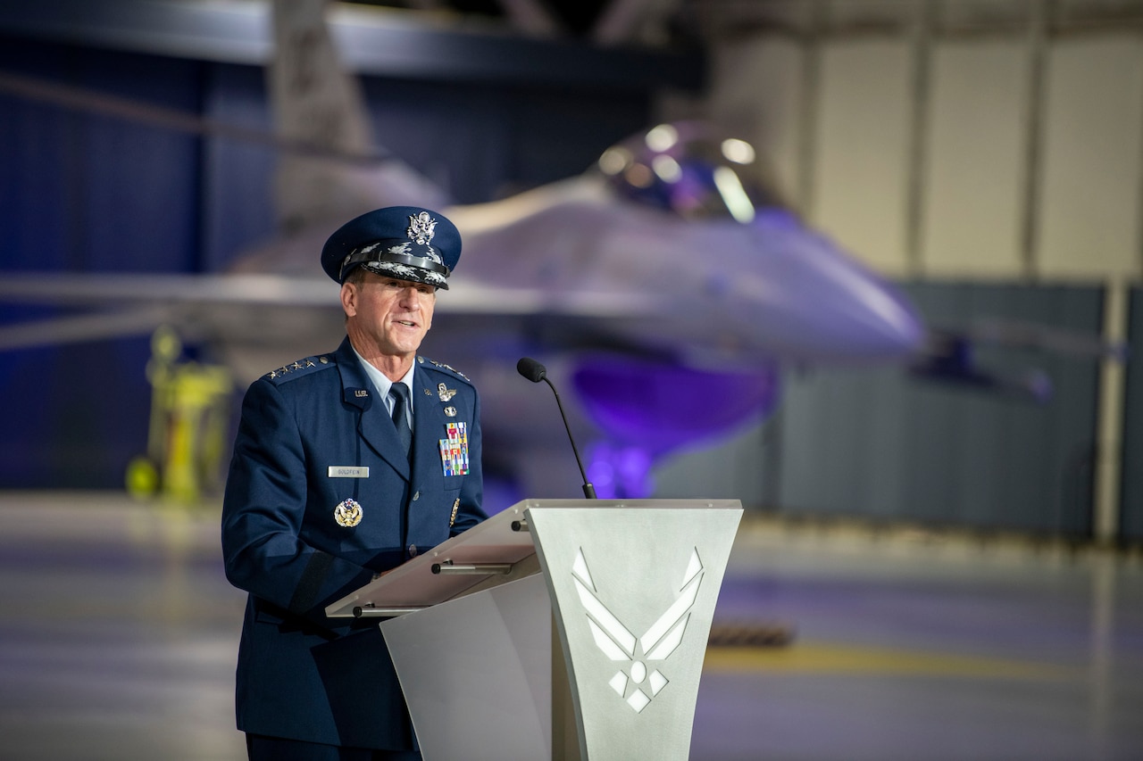 An Air Force general speaks at a lectern in an aircraft hangar with a jet in the background.
