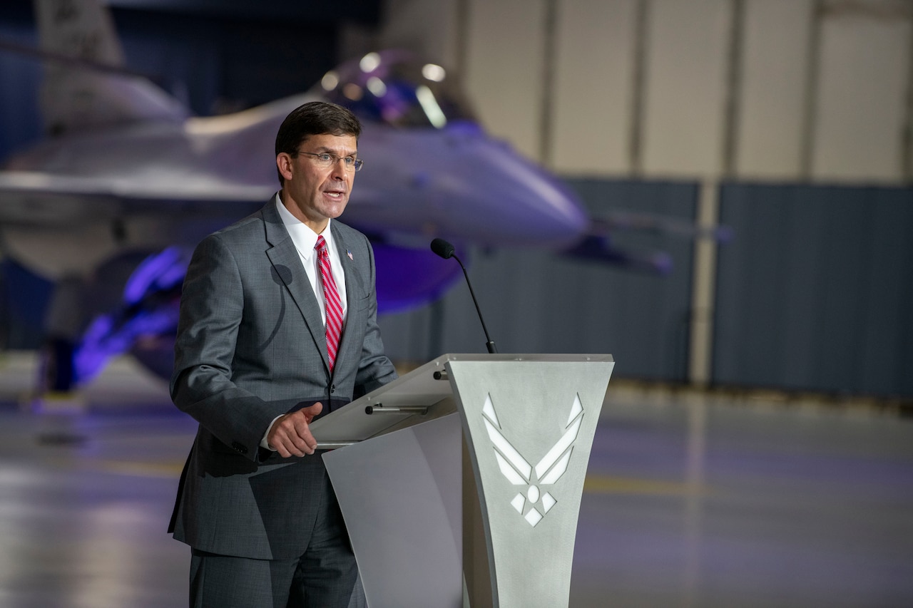 Man speaks at a lectern in an aircraft hangar with a jet in the background.