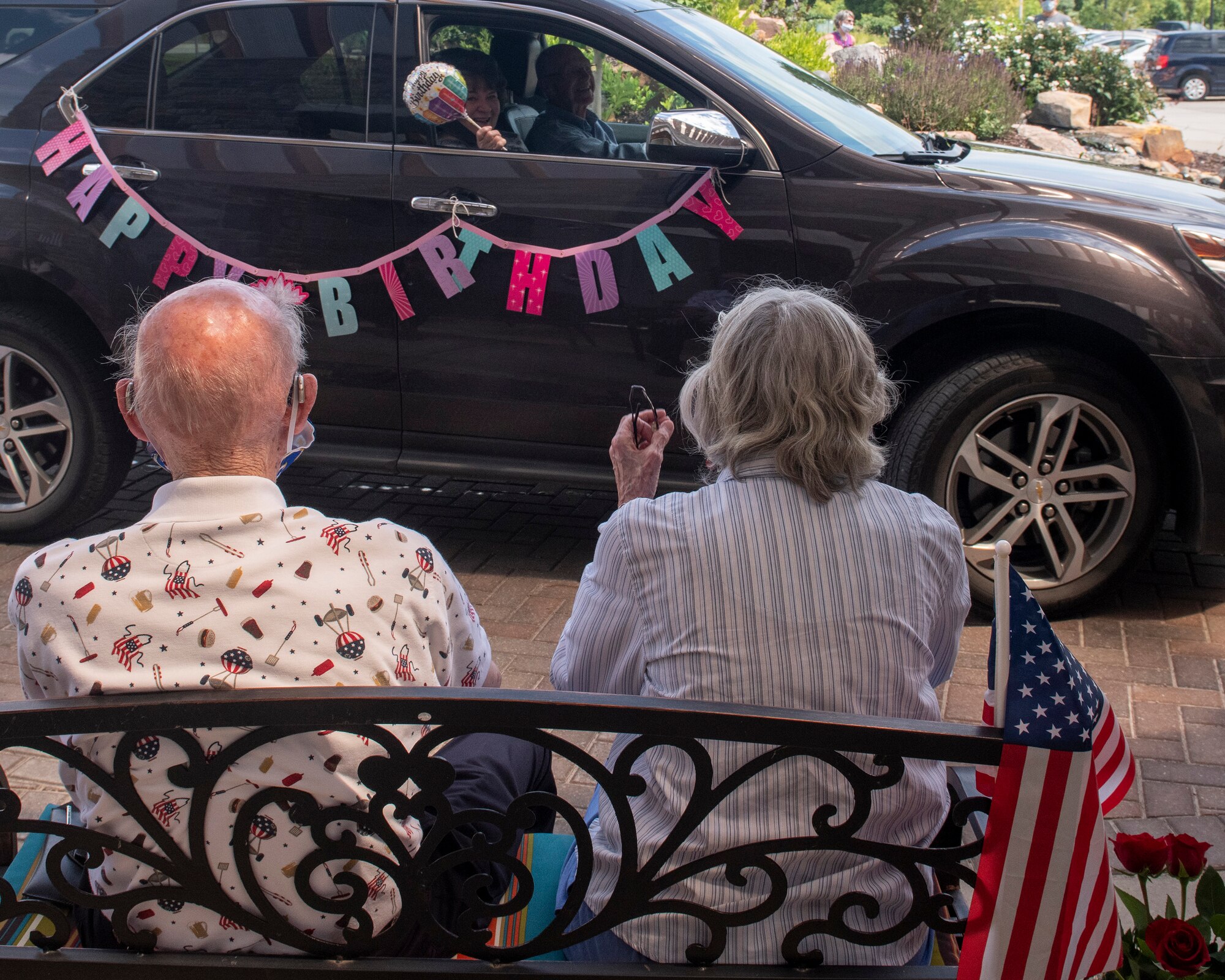 foreground of photo has retired CMSgt James McCoy and his wife Kathy sitting on a bench watching as a car with a Happy Birthday Banner pinned on it drives by