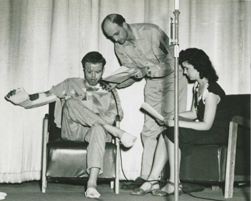 An injured man talks into a microphone another man is holding. A woman sits beside them.