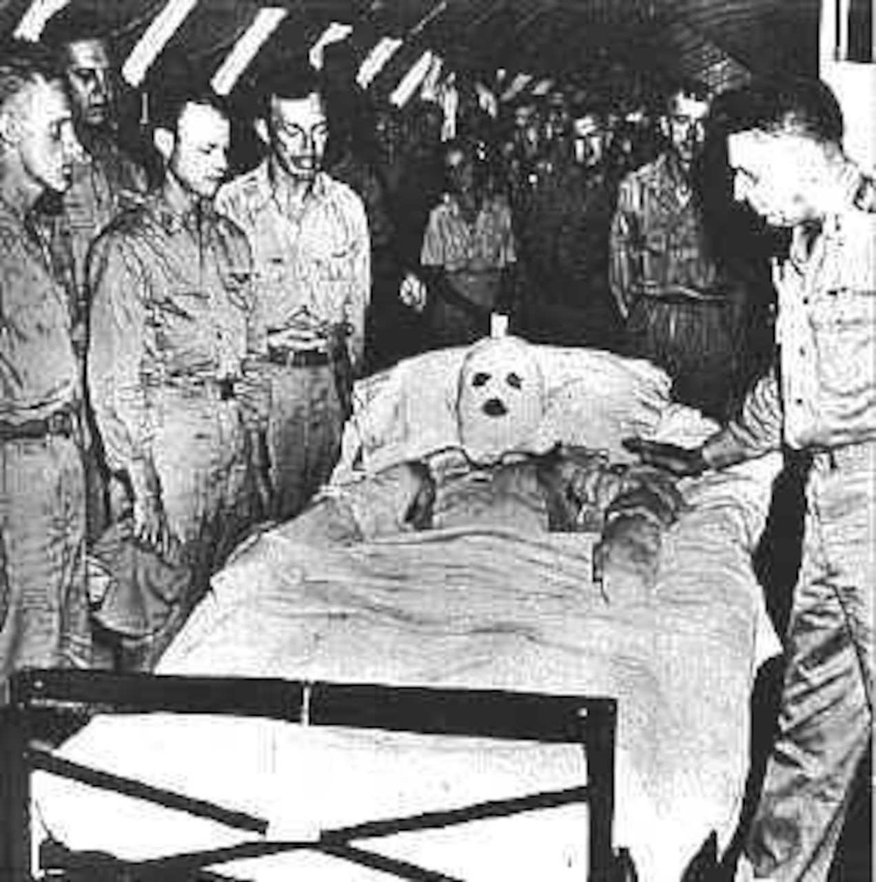 A man covered in bandages in a hospital bed is surrounded by soldiers.