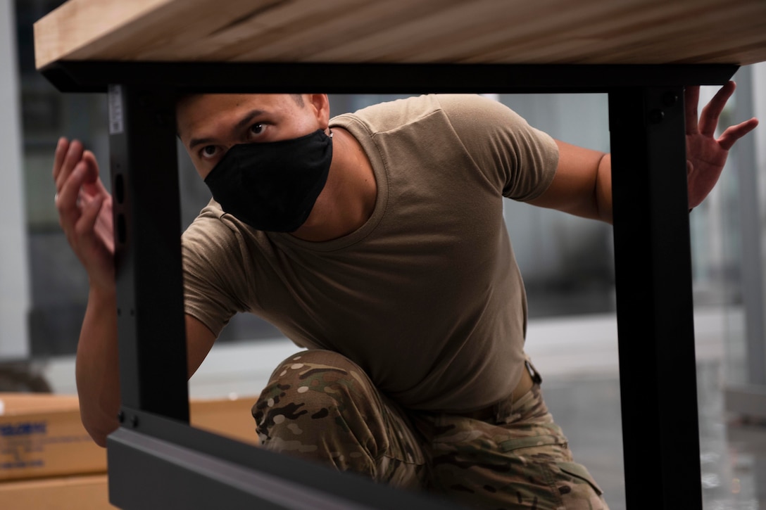 An airman wearing a face mask assembles a desk.