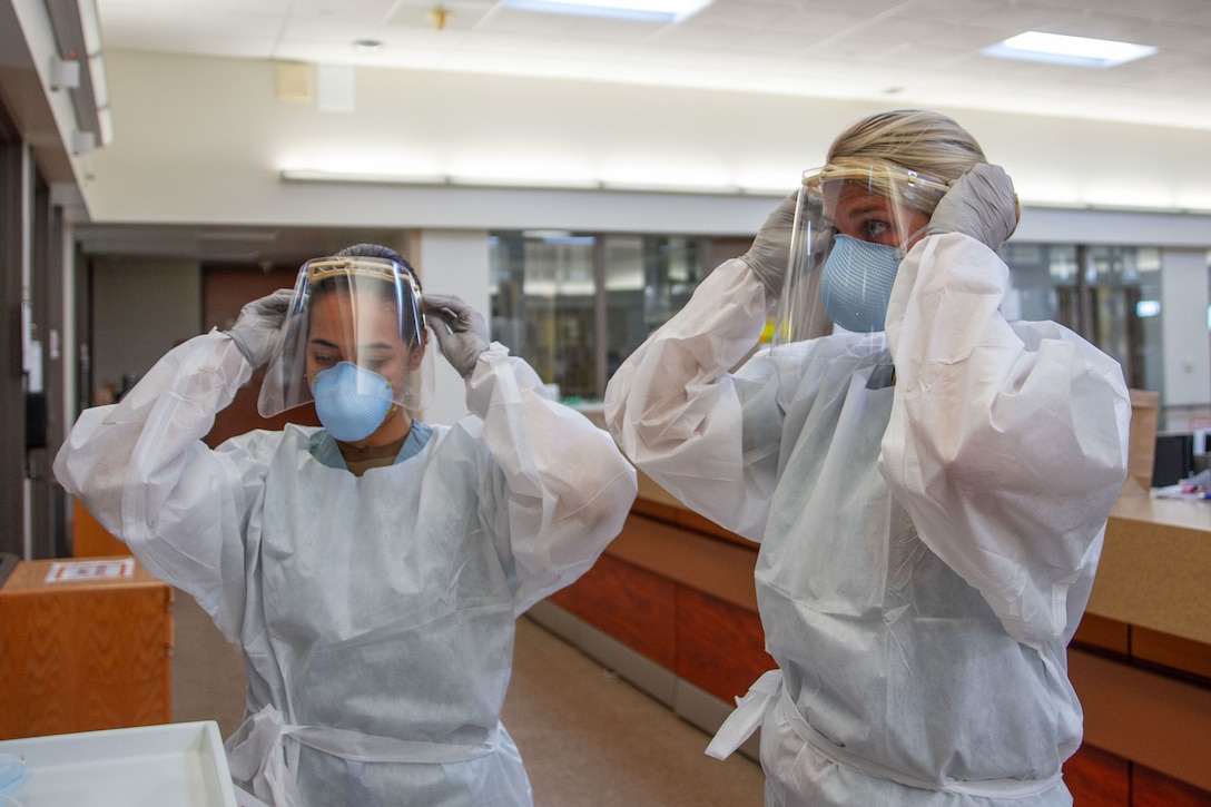 Two Navy sailors wearing personal protective equipment make sure to properly secure their face shields before entering a COVID-19 positive, non-critical patient’s room