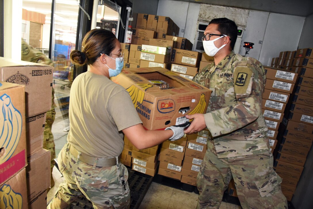 Two soldiers, one wearing a face mask and gloves and one wearing a face mask, carry a box of food that will be distributed at a local food bank.