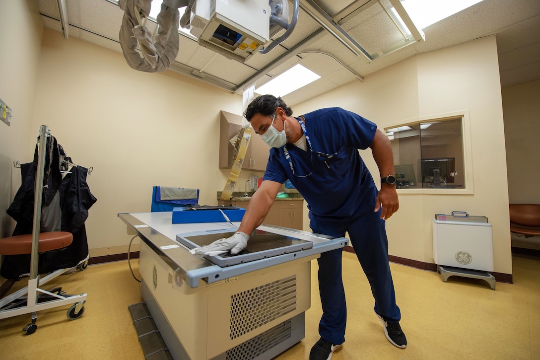 A radiology technician wearing a face mask and glove wipes down an X-ray machine.