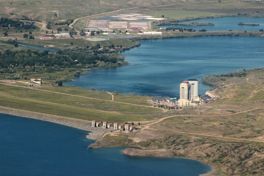 Looking downstream on the Fort Peck dam including the earthen embankment; the power house surge tanks to the right; the interpretive center on the left side of the embankment; and in the distance, at the top of the photo is the state fish hatchery.