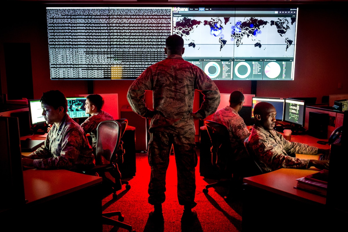 Military personnel sit at computer terminals in a room with a large screen.  One service member is standing.