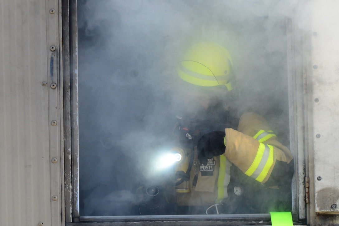 A man in a firefighting suit shines a light through some smoke.