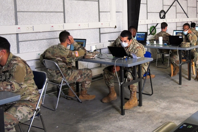 Soldiers sit at desks with laptops in front of them.
