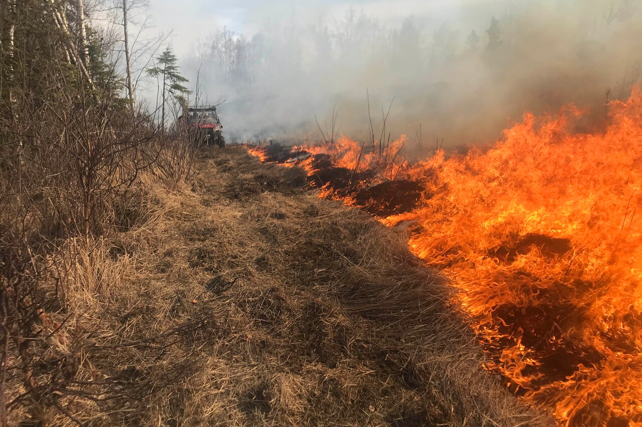 A fire burns along a line of brush. A firefighting vehicle is in the background.