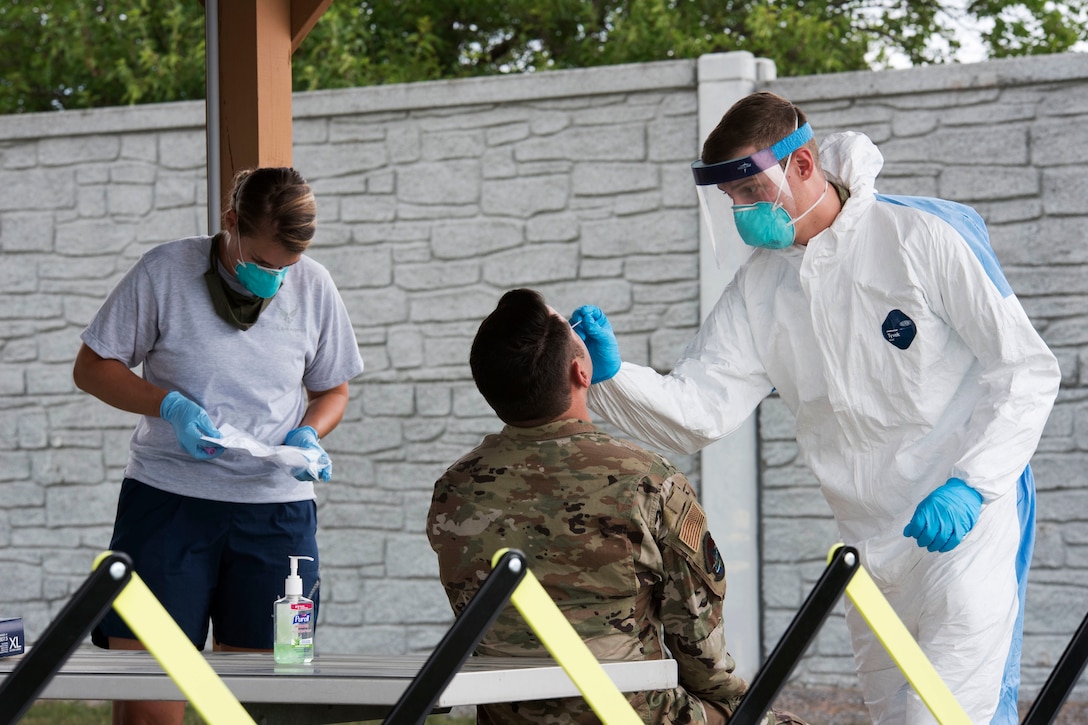 An airman wearing personal protective equipment administers a nasopharyngeal test to another airman while another medical tech wearing PPE assists.