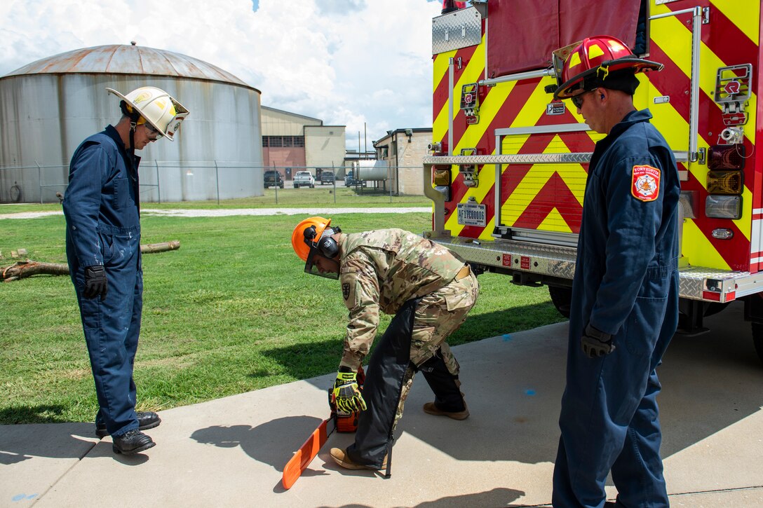 Two firefighters look on as a Soldier turns on a chainsaw.