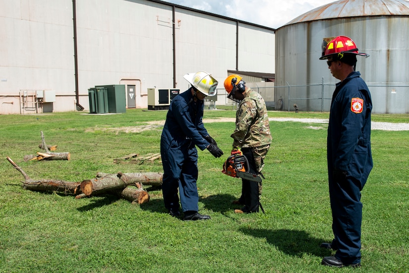 A firefighter looks on as another firefighter shows a Soldier how to use a chainsaw.