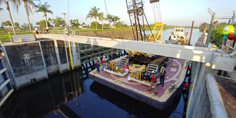 The crane operator begins the installation by setting a steel I-beam into the girder recess notch in the lock wall, starting with the upper or upstream end of the lock.