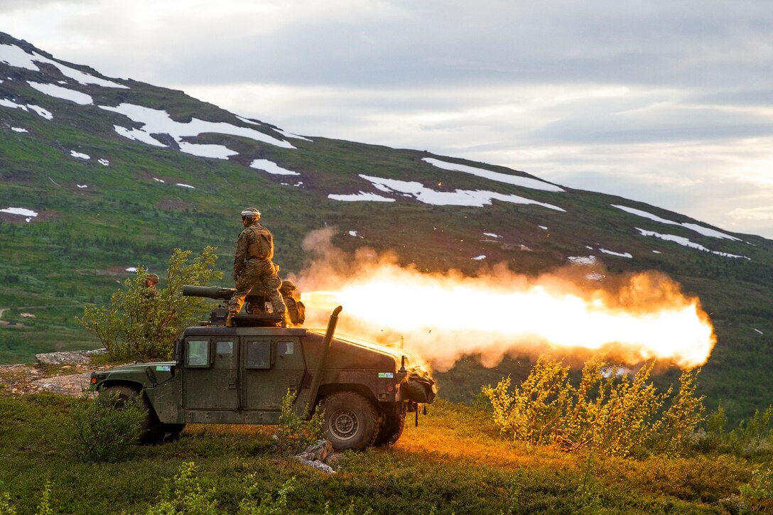 A Marine stands on a tank as a missile is fired near mountains.