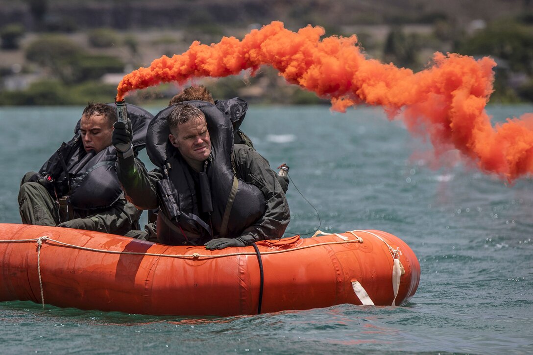 A Marine in an orange inflatable raft with other Marines holds up a flare that releases orange smoke.