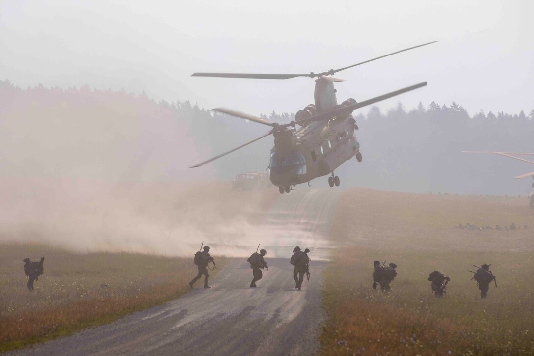 Soldiers run through a field underneath a military helicopter.