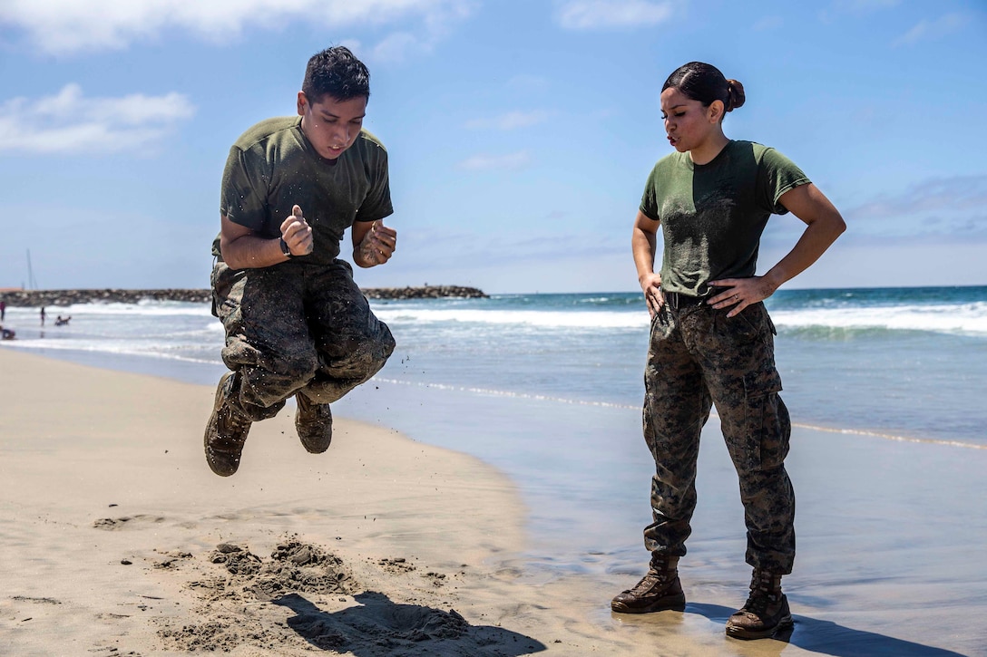 A Marine jumps next to another Marine on a beach.
