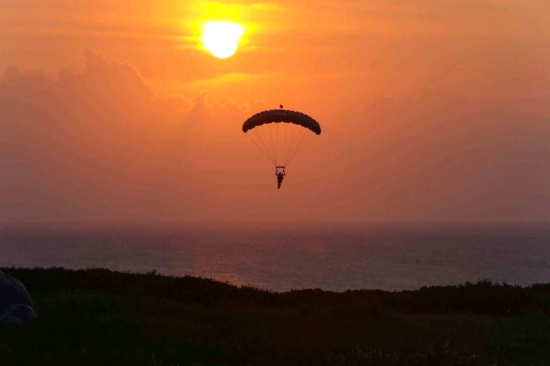 A Marine descends in the sky wearing a parachute as the sun shines in the background.