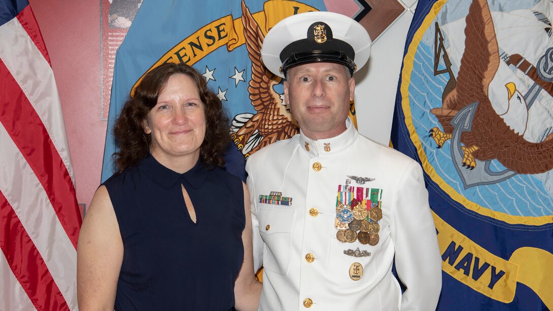 Man in white Navy dress uniform with medals and ribbons poses for a photo with a woman in a blue dress in front of a row of flags.