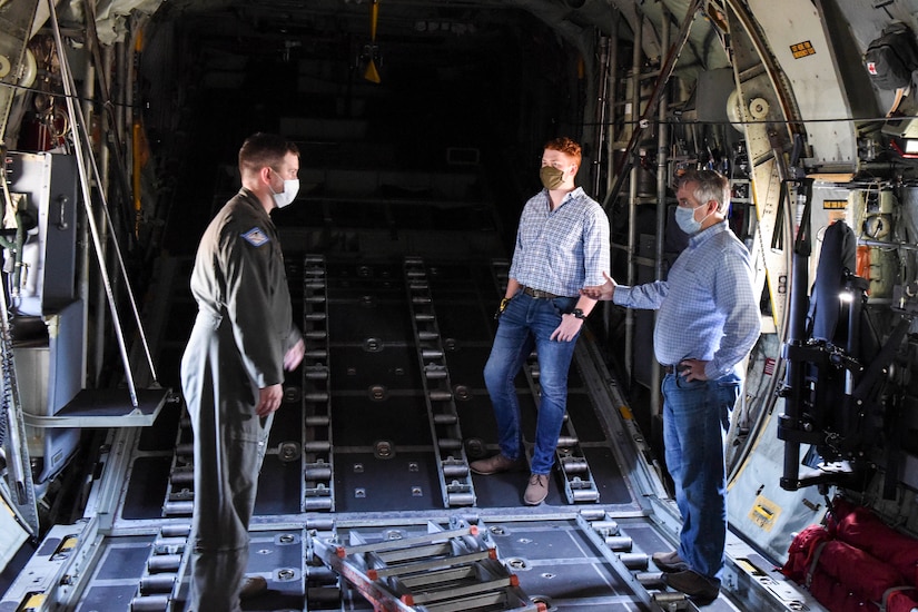 An officer wearing a flight suit talks with two civilians in the cargo bay of a transport aircraft. All are wearing face masks and practicing social distancing.