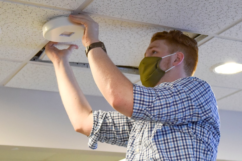 A man wearing a face mask installs an ultraviolet light on a ceiling.