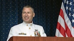 Man in white Navy dress uniform stands at a podium in front of the U.S. flag.