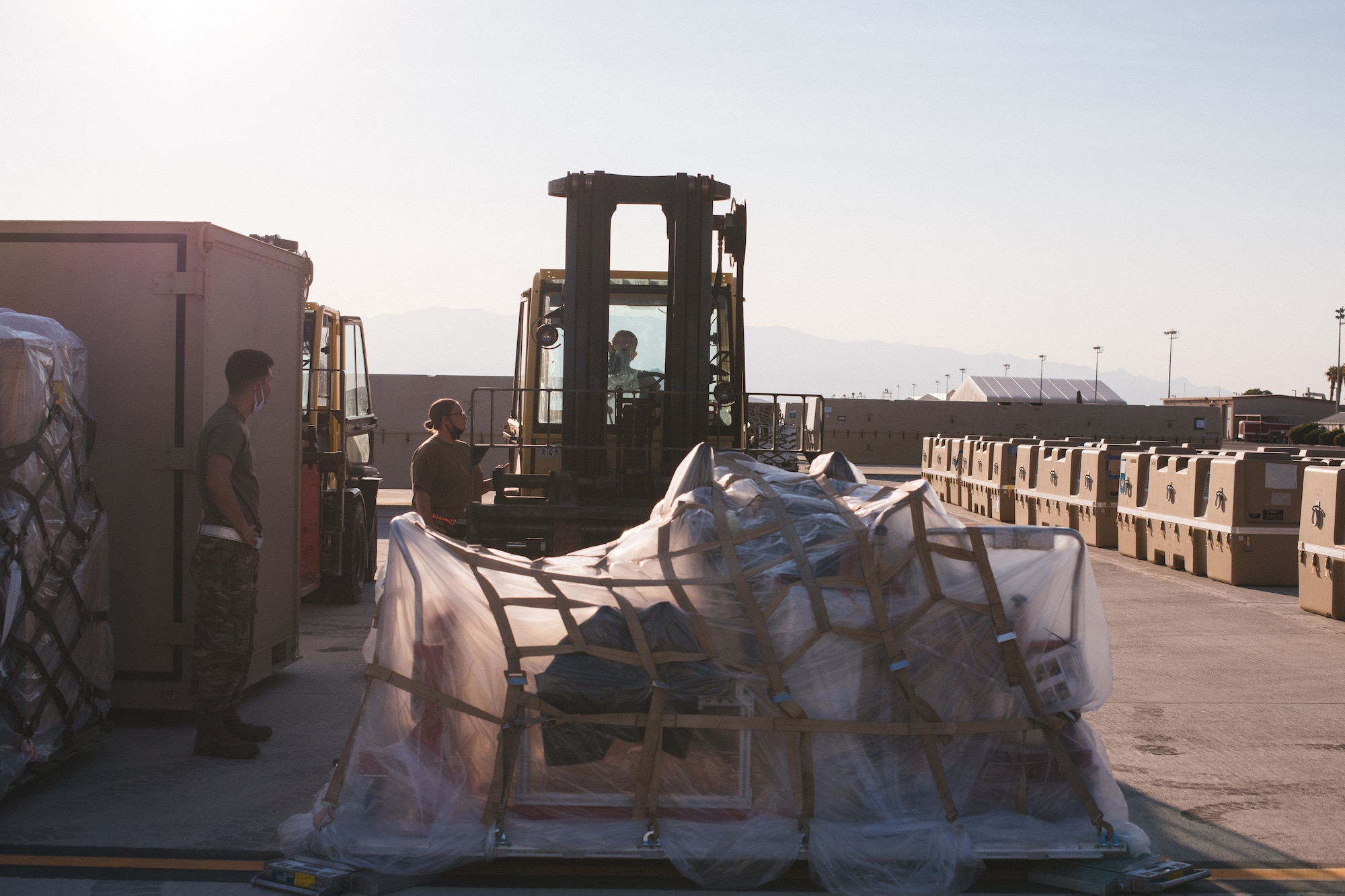 A piece of equipment in the foreground is lifted by a forklift as Airmen watch from the side.