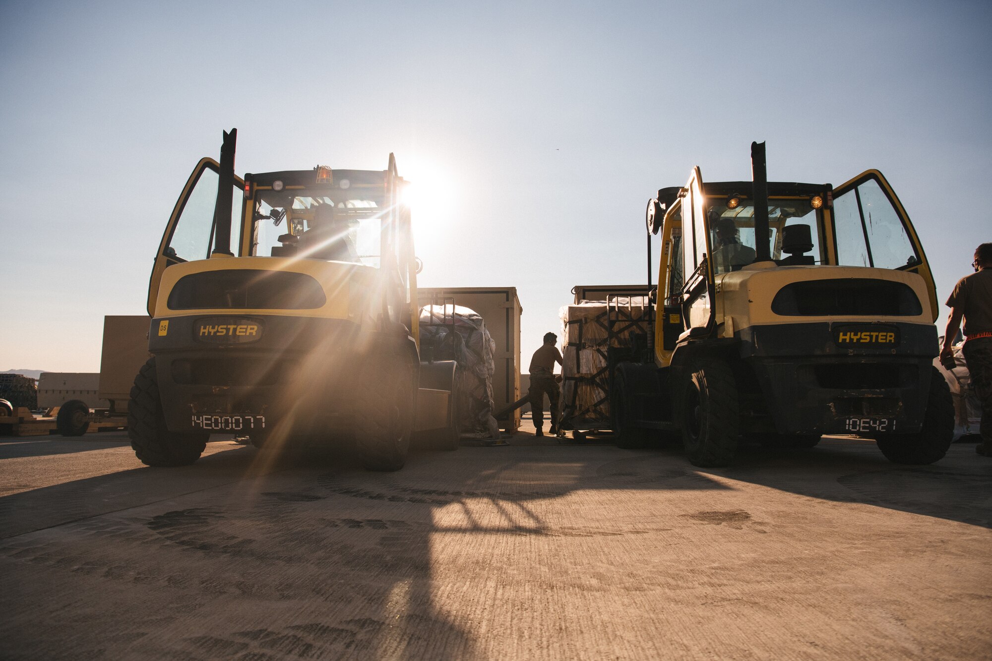 Two forklifts backlit by the sun are on either side of an Airmen in the center of the photo.