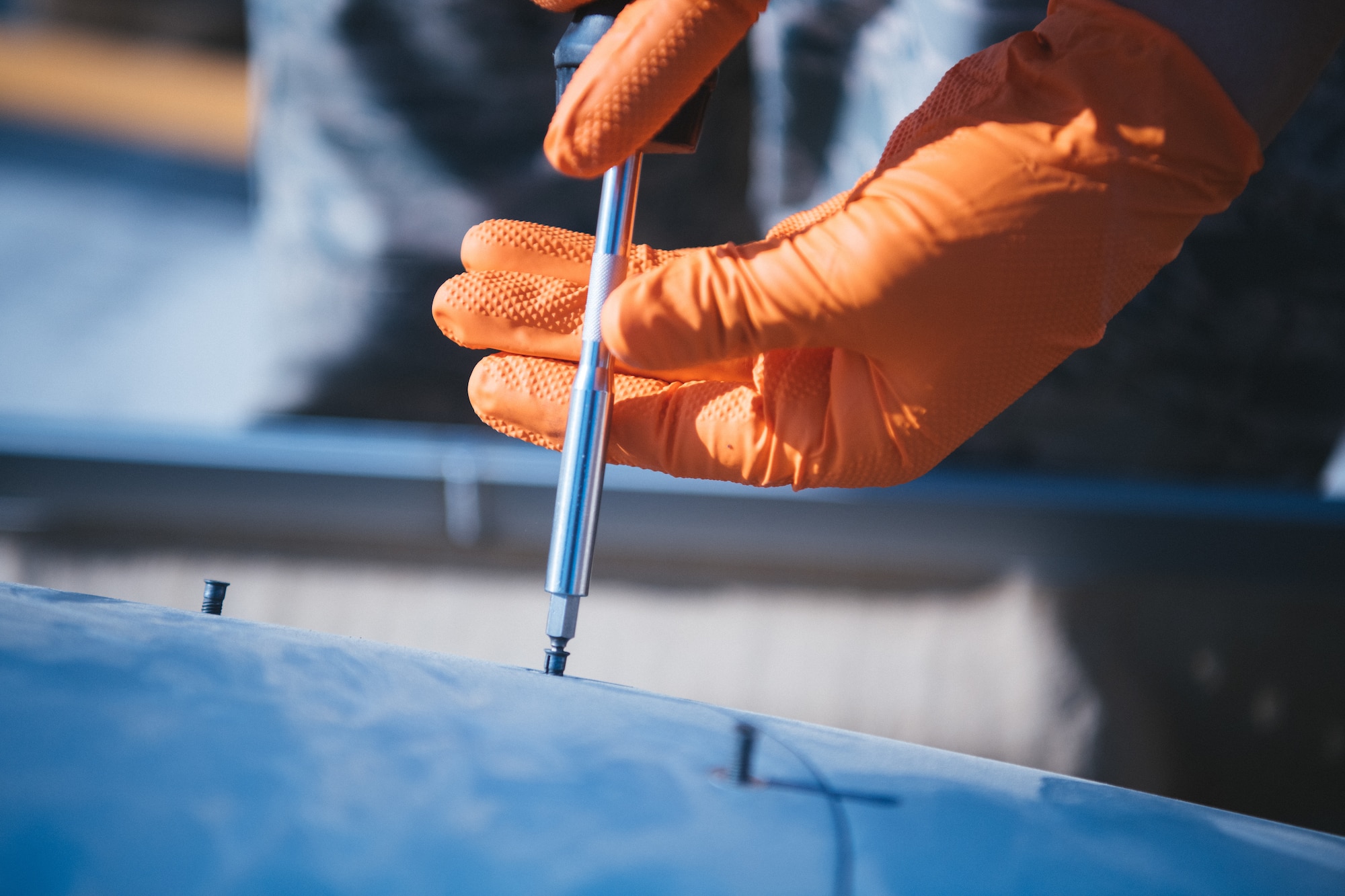 Hands wearing orange gloves unscrew a fuel tank panel.