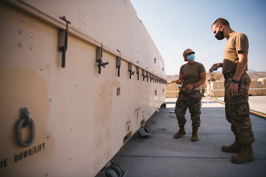 Two Airmen are dwarfed by the size of a MQ-9 Reaper connex as they look at it.