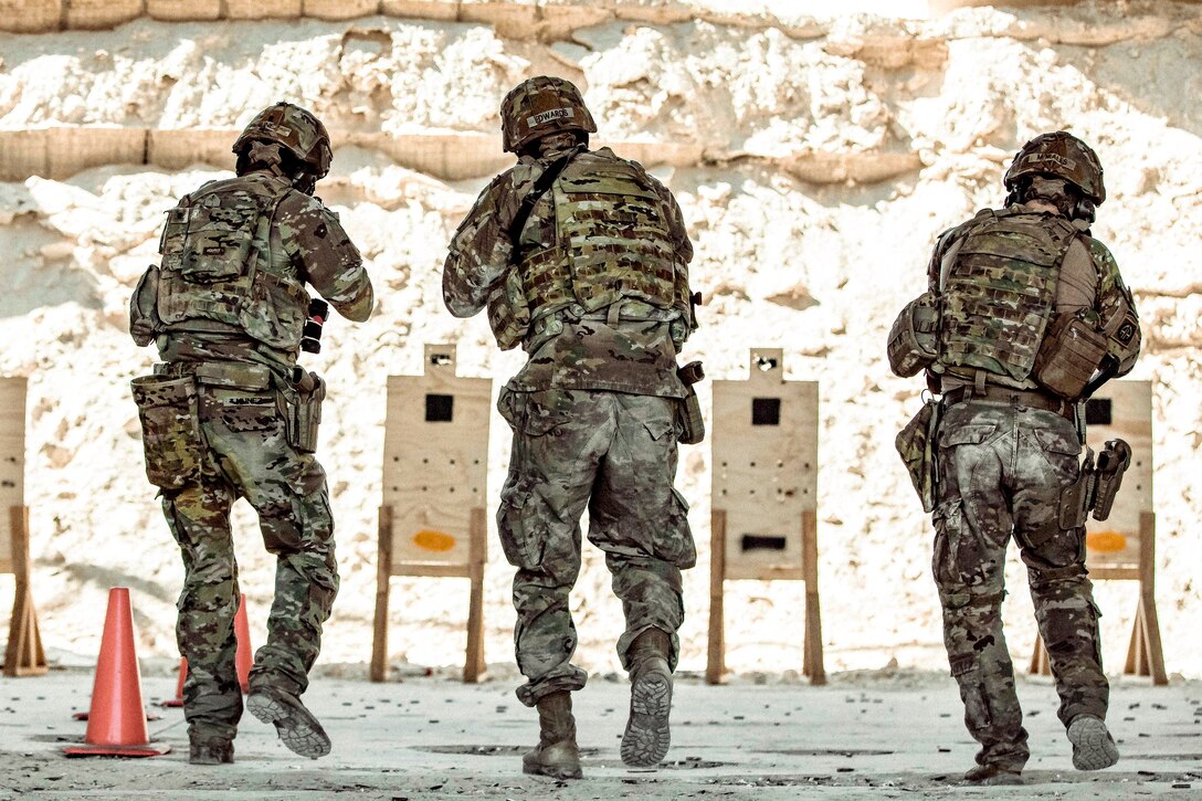 Three soldiers, shown from behind, move toward targets at an outdoor firing range.