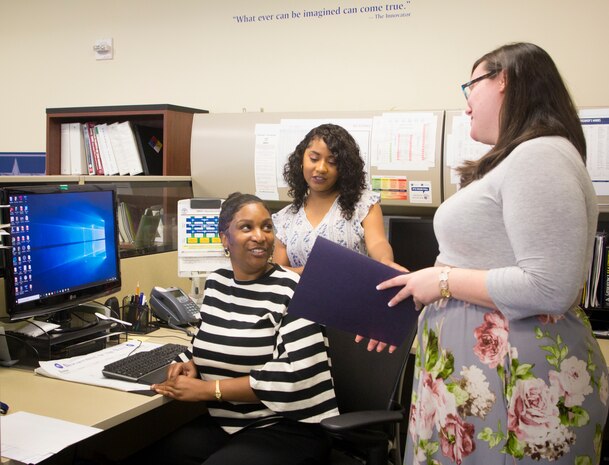 Timberlain Woodruff, seated, talks with coworkers Amber Cooke, middle , and Tonya MacLunny. Woodruff, a Navy Warfare Development Command (NWDC) management analyst, is one of eight people selected to receive the Judith C. Gilliom Award for Outstanding Workforce Recruitment Program participants. NWDC develops and integrates innovative solutions to complex naval warfare challenges to enhance current and future warfighting capabilities. (U.S. Navy photo by Ian Delossantos)