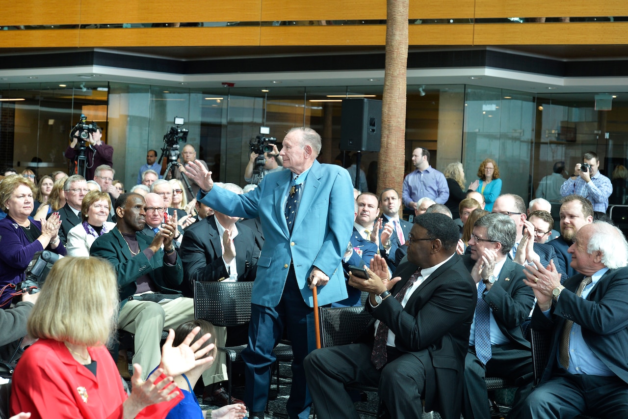 An elderly man with a cane stands in a crowd and waves as they applaud.