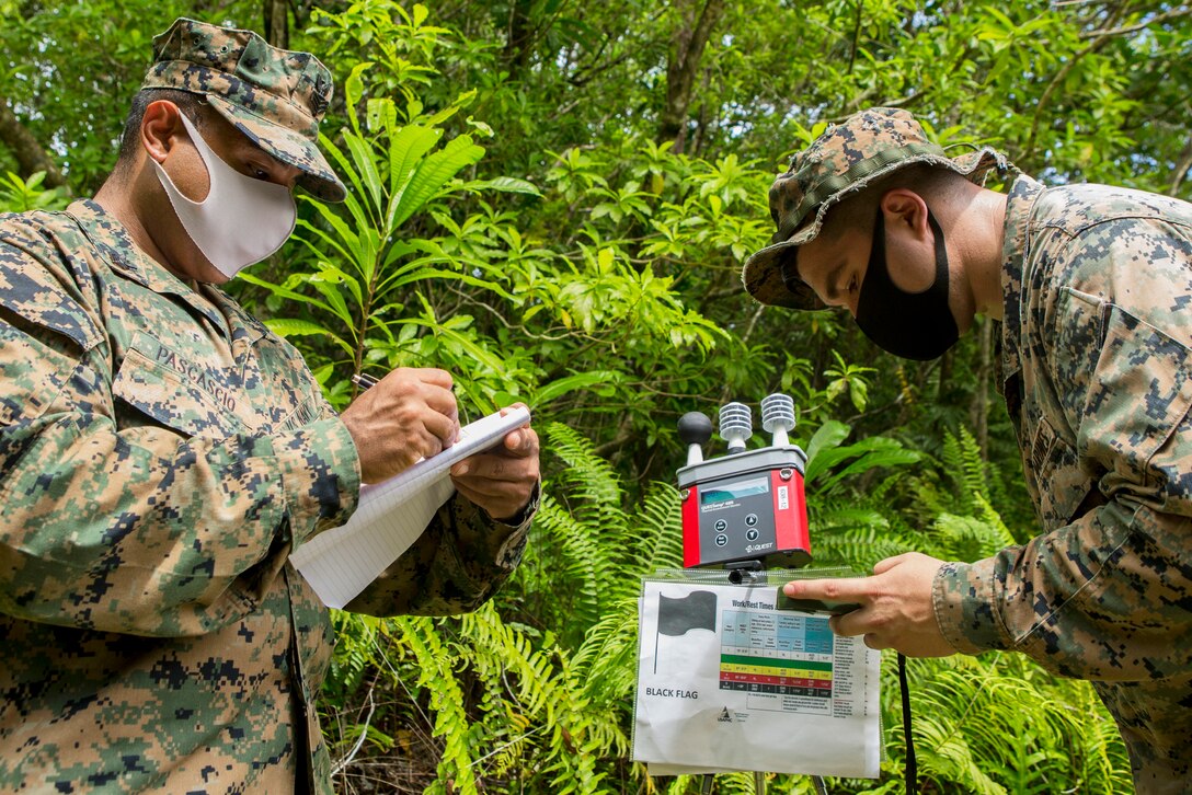 A Marine and a sailor, wearing face masks, observe and annotate climate data using a wet bulb globe thermometer and a Kestrel handheld weather meter.