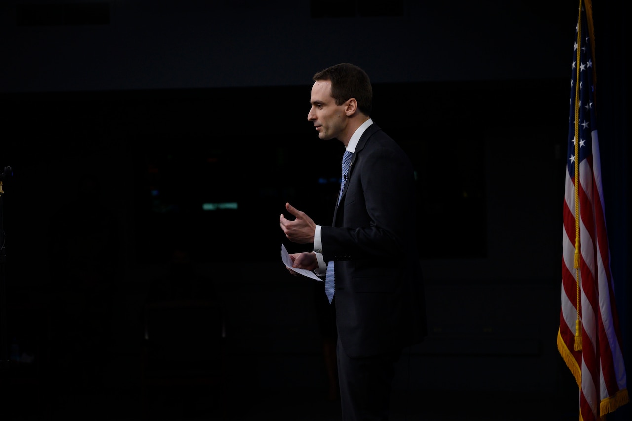 A man stands in a dark room facing an audience. A U.S. flag is behind him.