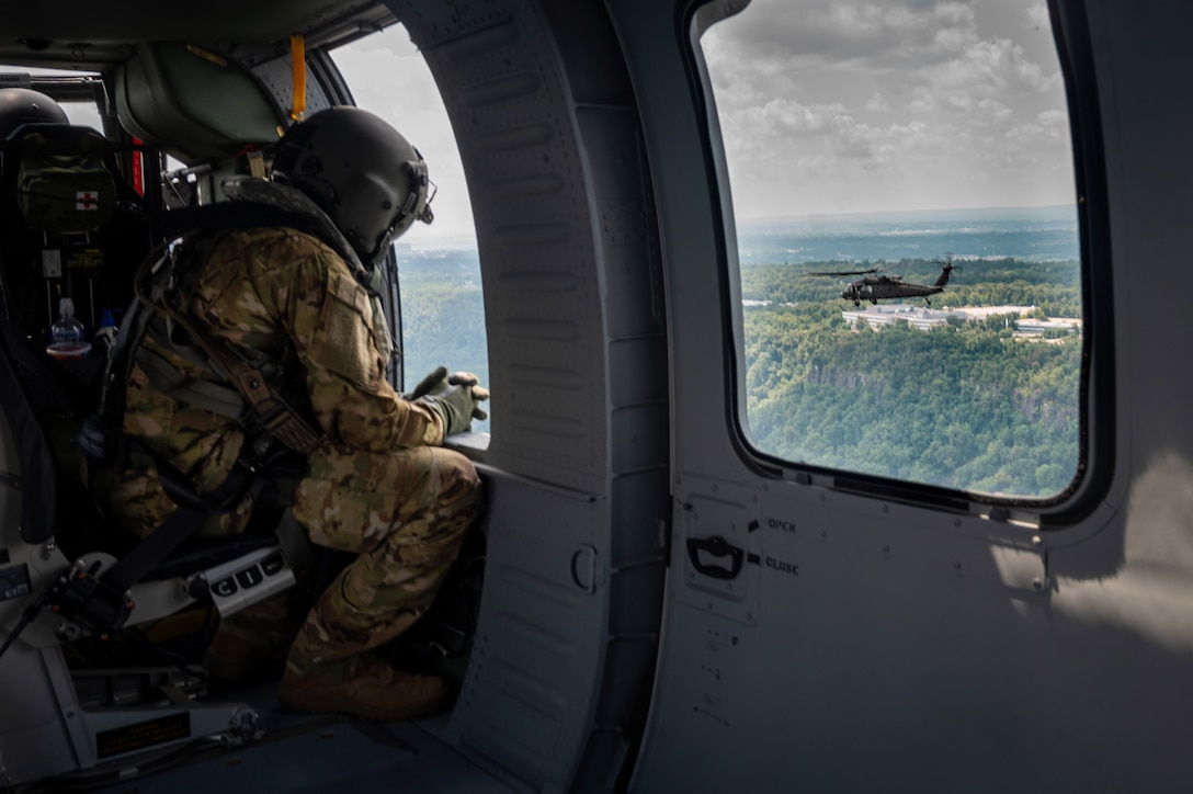 A Guardsman looks out of a helicopter window while another helicopter passes by.