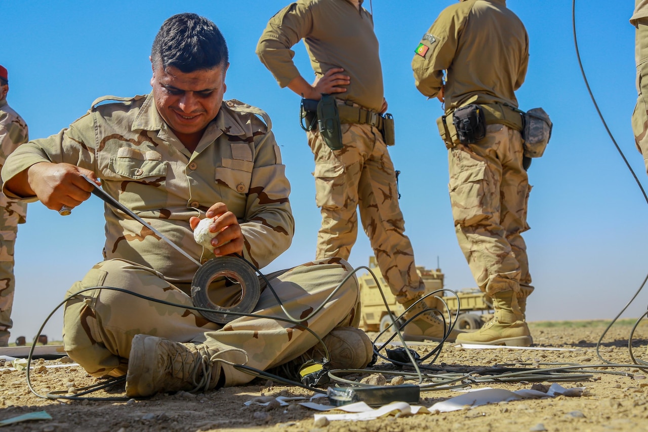 A man in a military uniform unrolls tape from a spool. Other military personnel stand nearby.