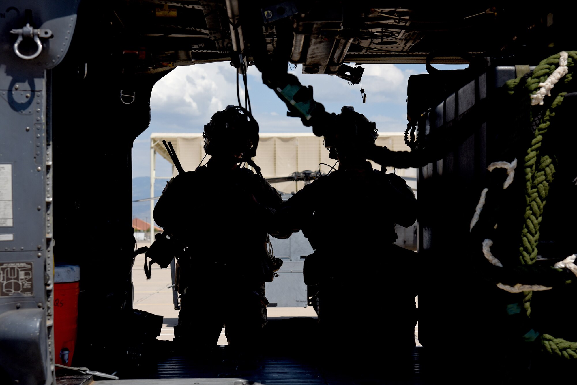 Photo of two Air Force Airmen casting a silhouette behind the side door of an HH-60G Pavehawk on a military flightline