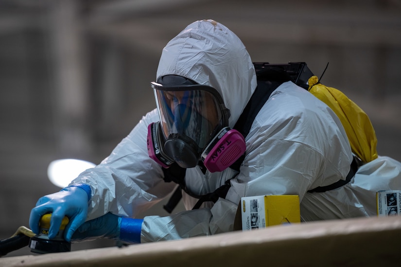 An Airman wears a Tyvek coverall and a personal cooling system as he sands a panel on an F-22 Raptor's wing.