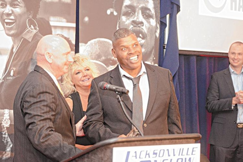 Man smiles at lectern while standing in front of a woman and beside a man.