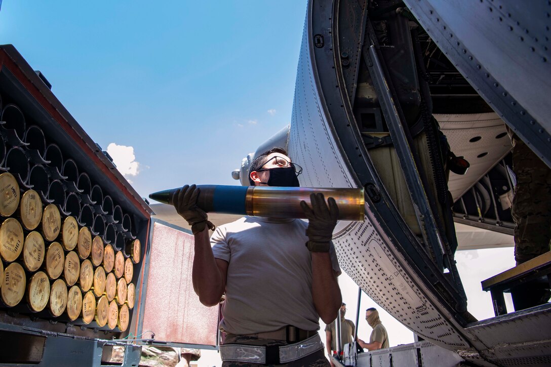 An airman wearing protective gear holds ammunition near an aircraft.