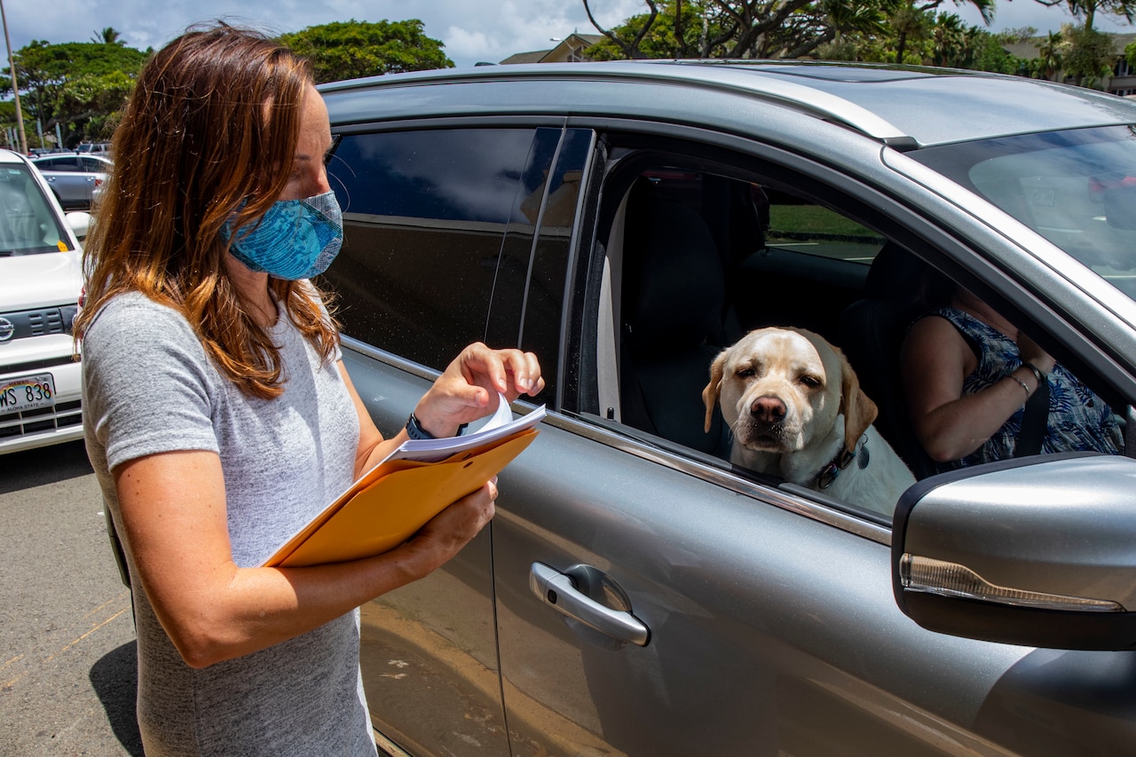 A masked woman, holding papers, talks to a driver in a car.