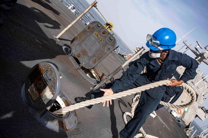 A sailor in a blue hard hat pulls a rope from inside a ship.