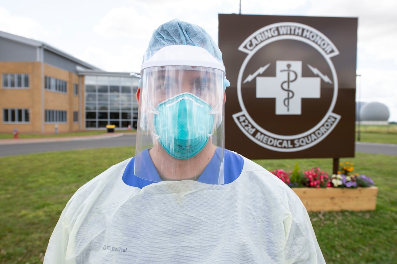 An airman in medical protective gear poses in front of a medical center sign.