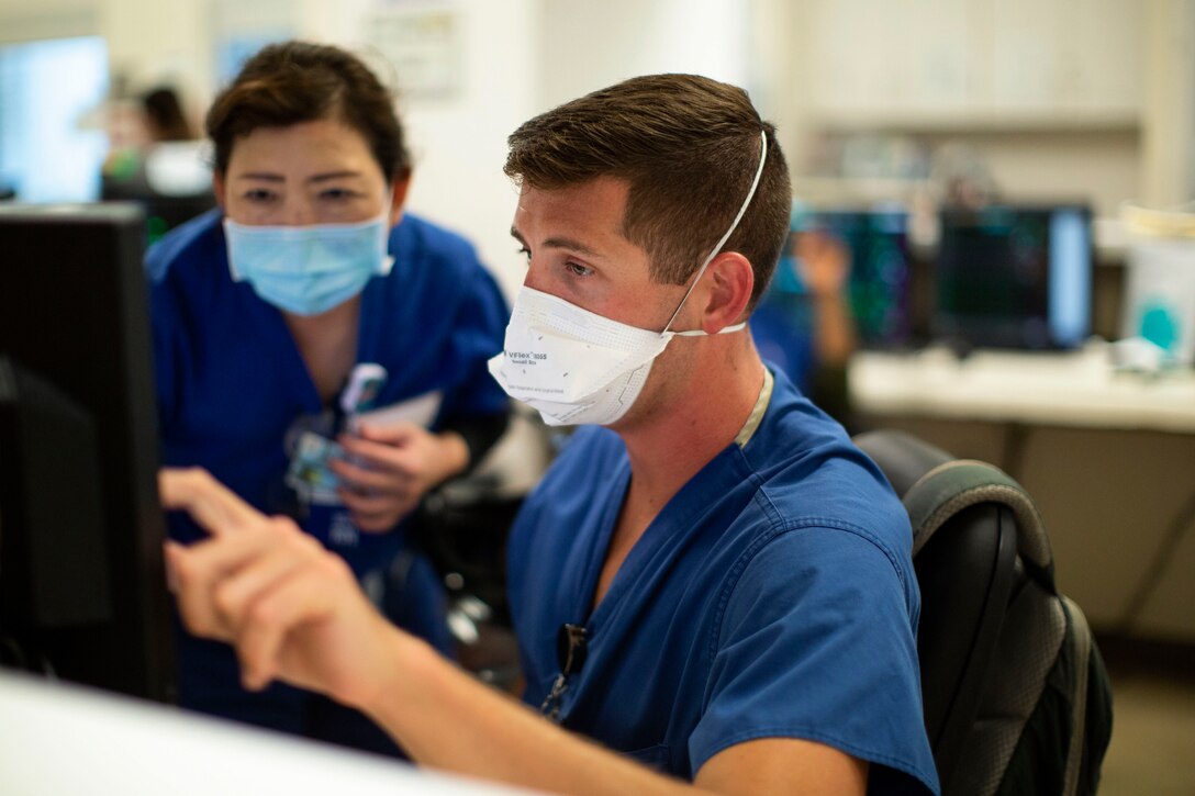 An airman in protective gear looks at a computer screen with another service member.